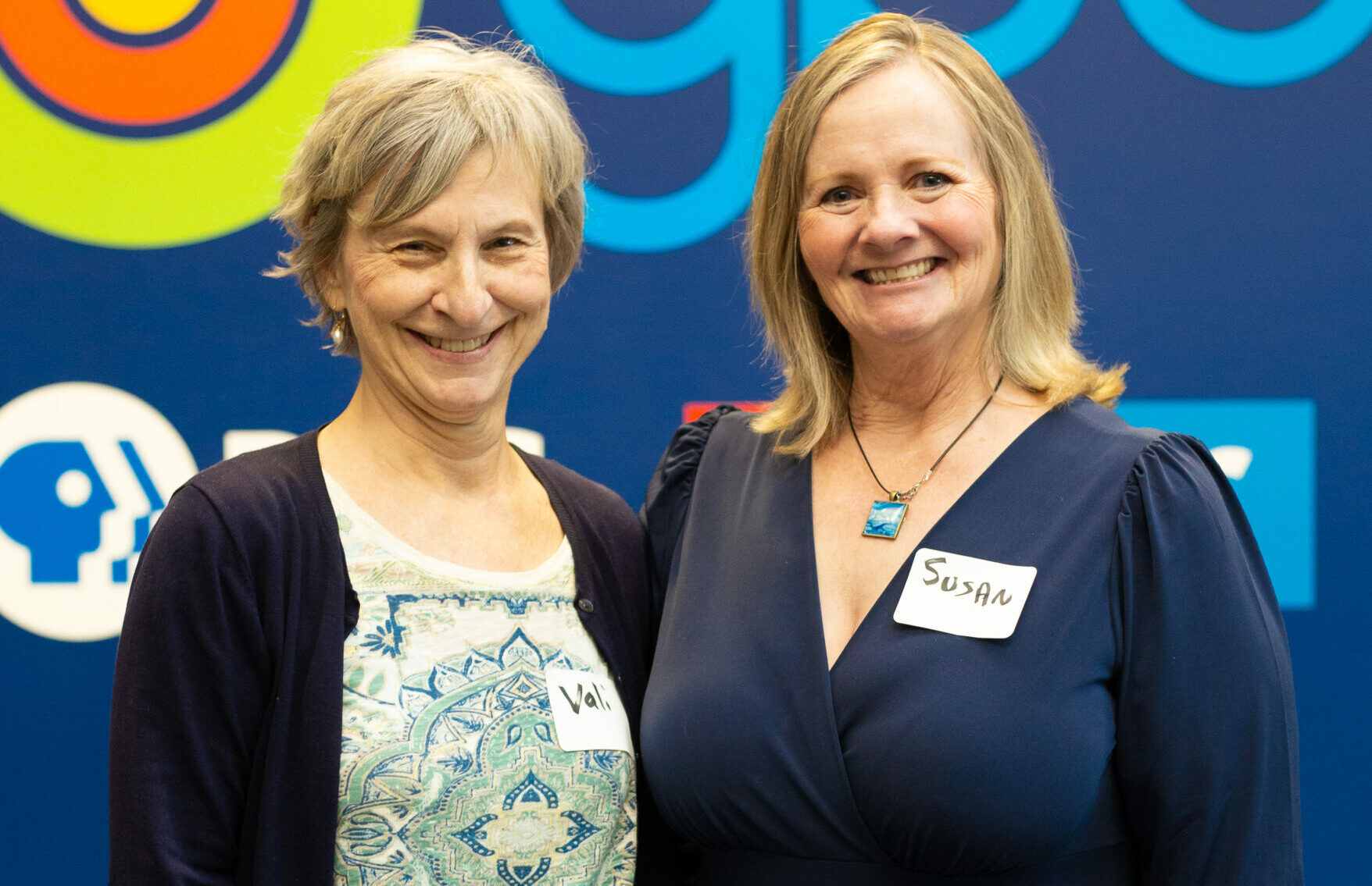 This image features two smiling women standing side by side in front of a dark blue backdrop with colorful logos, including those of GPB (Georgia Public Broadcasting) and PBS. The woman on the left, named Val (as shown on her name tag), has short gray hair and is wearing a navy cardigan over a light shirt with an intricate, mandala-like pattern. She is also wearing high-waisted blue jeans and holding a pencil in her right hand. The woman on the right, named Susan (according to her name tag), has blonde hair and is dressed in a deep navy blue dress with a v-neckline. She is wearing a turquoise pendant necklace and a watch on her left wrist. A tattoo is partially visible on her right forearm. Both women appear warm and friendly, standing closely together as if posing for a commemorative photo at an event or gathering.