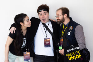 Three attendees at VO Atlanta 2025 stand closely together smiling and making playful faces. The person in the middle has a surprised expression, with one arm around each of the others. All wear conference badges, and the person on the right carries a SAG-AFTRA tote bag with "#AdsGoUnion" written on it.