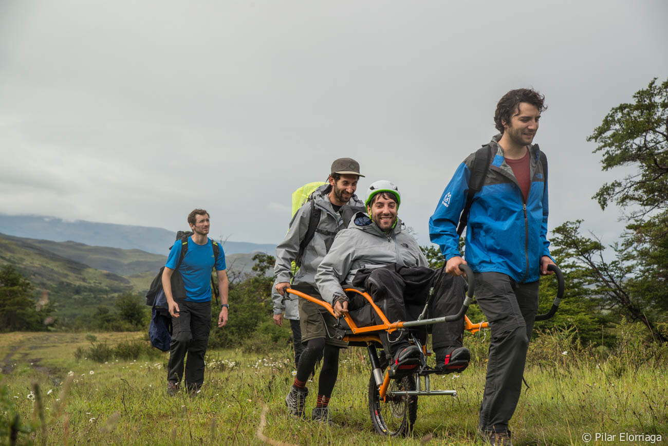Four hikers are outdoors in a grassy mountain landscape. Two men are carrying another man with a disability in an adaptive hiking chair with a single wheel, while a fourth man walks beside them with a backpack. Everyone appears to be enjoying the hike, smiling and working together as a team.