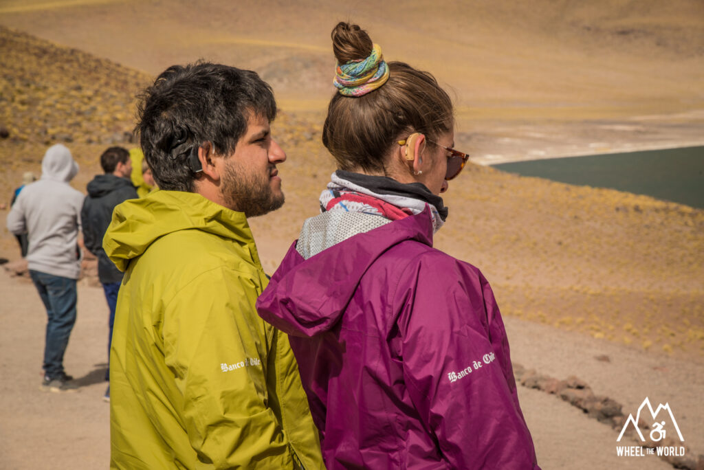 Two travelers wearing bright outdoor jackets stand side by side, looking out over a desert landscape with a lake in the distance. Both are using hearing devices, representing accessibility and inclusion in outdoor adventures.