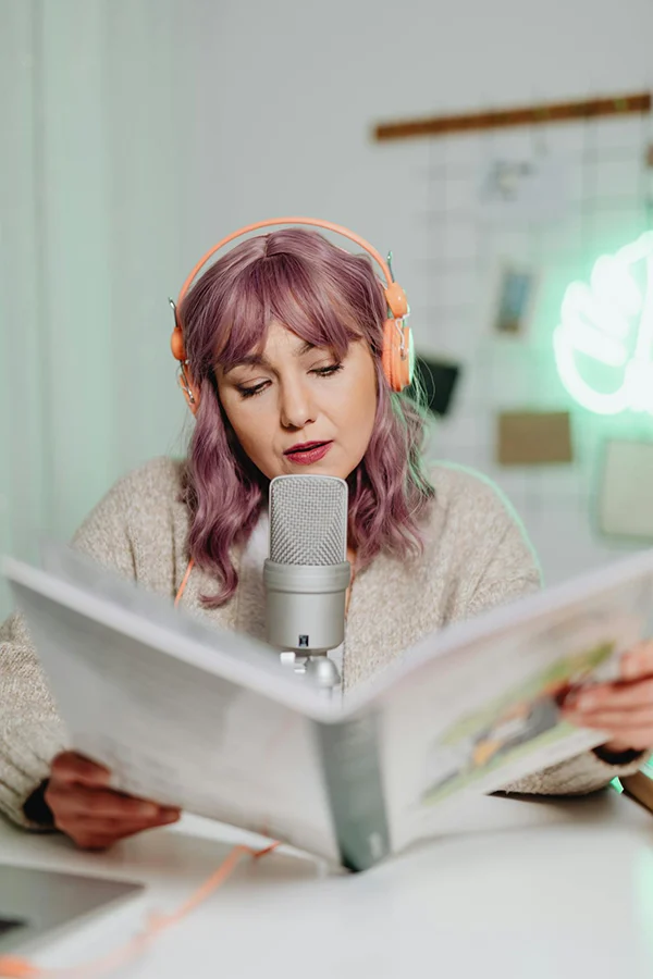 A woman with purple hair and orange headphones reads a newspaper into a studio microphone.