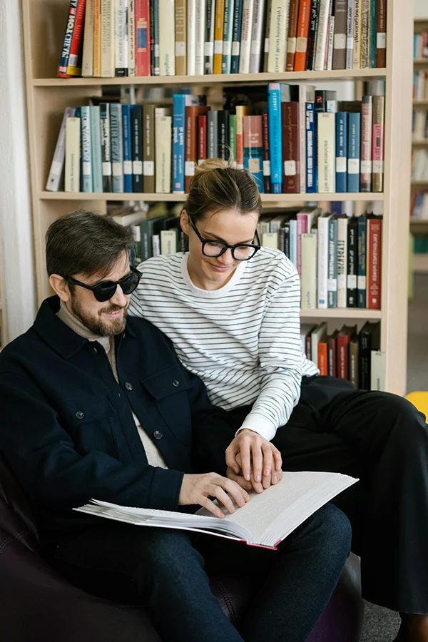 A woman guides a man wearing dark glasses as they read a Braille book together in a library.