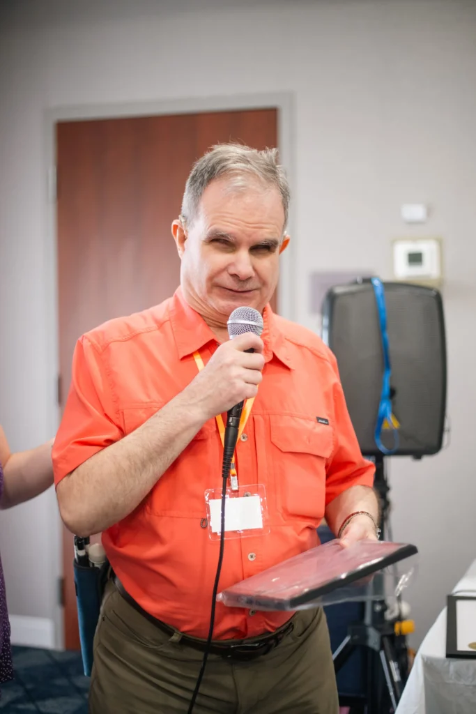 A Blind Man In An Orange Shirt Speaks Into A Microphone While Holding A Book, Addressing An Audience.