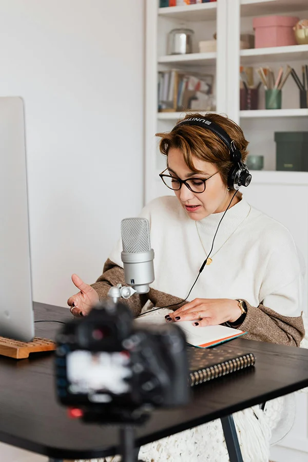A woman wearing headphones speaks into a microphone while reading notes at a desk, with a camera recording her.