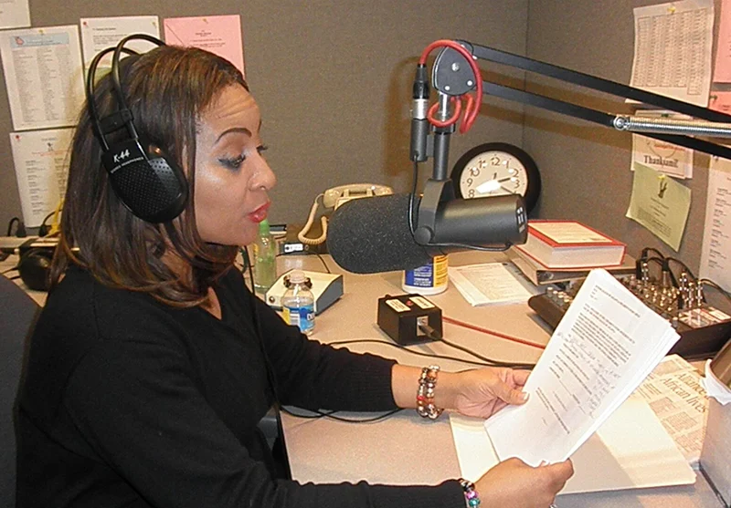 Woman wearing headphones reads from a script while speaking into a studio microphone in a radio recording booth.